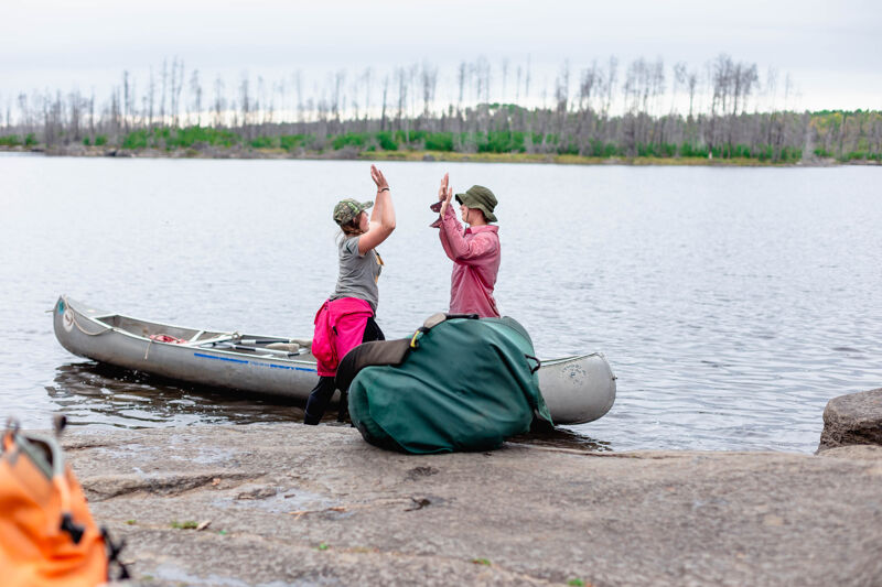 Two people are giving each other a high five next to a canoe on the shore of a lake. One person is wearing a pink skirt and a hat, and the other is wearing a hat and a pink shirt. There is a large green bag next to the canoe. The lake is calm and there are trees in the background.